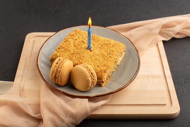 Uma fatia de bolo de vista frontal saborosa e cozida dentro da placa com vela. Macarons na mesa de madeira e bolo escuro de biscoito de açúcar doce