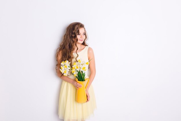 Uma foto de estúdio de uma garota de pé e segurando um vaso amarelo com flores. A garota tem cabelos louros compridos e encaracolados e usa um vestido amarelo desolado. A menina sorri e parece tímida.