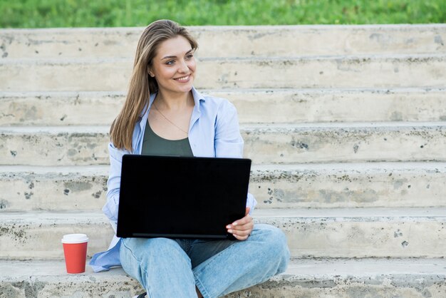 Uma garota caucasiana feliz e atraente está sentada com um laptop nas escadas. trabalho à distância. conceito freelance