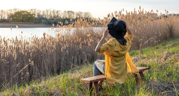 Uma garota com um chapéu e estilo casual se senta em um banco perto do lago ao pôr do sol.