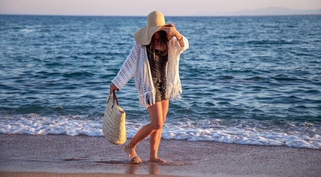 Uma garota com um grande chapéu com uma bolsa de vime caminha à beira-mar. Conceito de férias de verão.