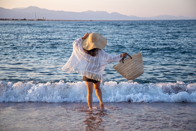 Uma garota com um grande chapéu e uma bolsa de vime caminha ao longo da costa do mar. Conceito de férias de verão.
