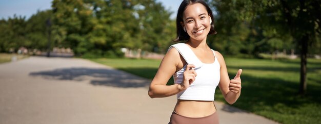 Foto grátis uma garota de fitness sorridente mostra o polegar para cima, fica com uma toalha depois de um bom treino no parque, correndo e olhando