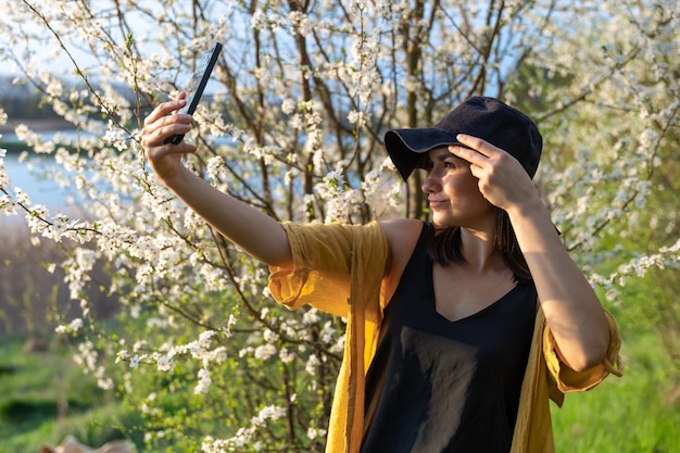 Uma garota estilosa com um chapéu fazendo uma selfie ao pôr do sol perto de árvores floridas na floresta