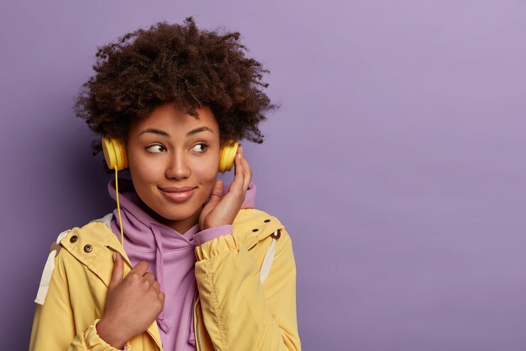 Foto grátis uma garota hipster de pele muito escura ouve um audiolivro em fones de ouvido, focada ao lado com uma expressão sonhadora e usa roupas casuais
