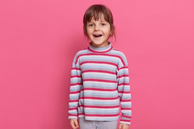 Uma garota pré-escolar feliz e animada usando uma camisa listrada estilo casual posando isolada sobre uma parede rosa