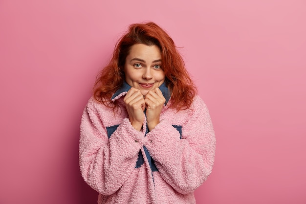 Uma garota ruiva bem satisfeita se sente confortável usando um novo casaco quente rosa, olha diretamente para a câmera, tem covinhas nas bochechas