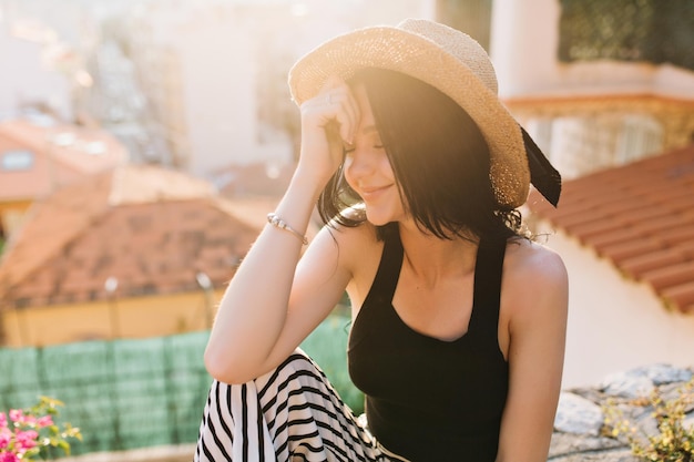 Uma garota sonhadora de cabelos escuros usando uma elegante pulseira de prata sentada com os olhos fechados, aproveitando o sol. adorável jovem morena de chapéu e top preto tomando banho de sol no telhado na manhã ensolarada.
