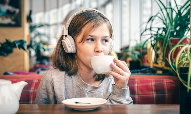 Uma garotinha em fones de ouvido em um café com uma xícara de chá