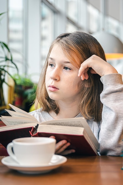 Uma garotinha está lendo um livro enquanto está sentado em um café com uma xícara de chá