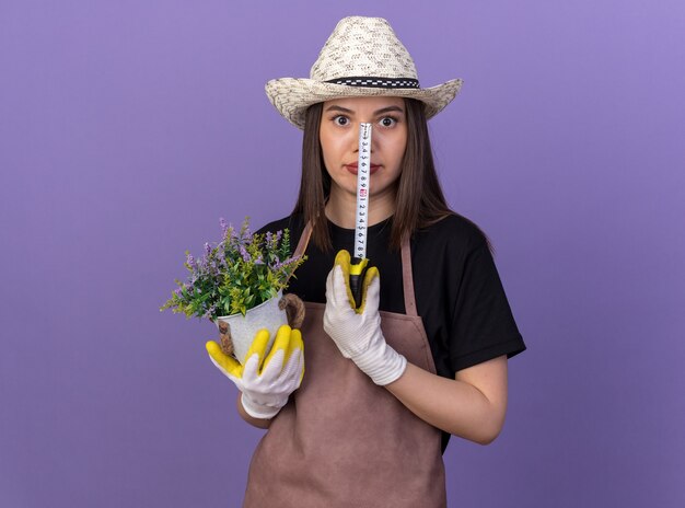 Uma jardineira caucasiana bonita e surpresa com um chapéu de jardinagem segurando um vaso de flores e uma fita métrica