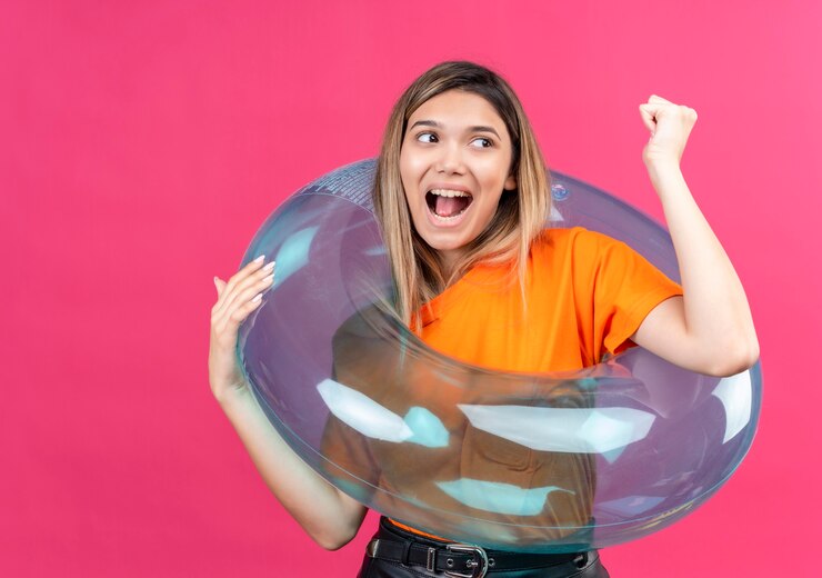 Foto grátis uma jovem adorável e feliz com uma camiseta laranja levantando o punho cerrado enquanto está de pé em um anel inflável em uma parede rosa
