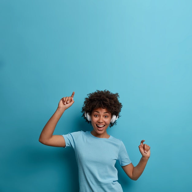 Foto grátis uma jovem afro-americana encantada positiva aprecia a melodia da música, usa fones de ouvido sem fio, se move com o ritmo da música, tem um humor alegre, isolada na parede azul, copie o espaço acima. descanso, estilo de vida