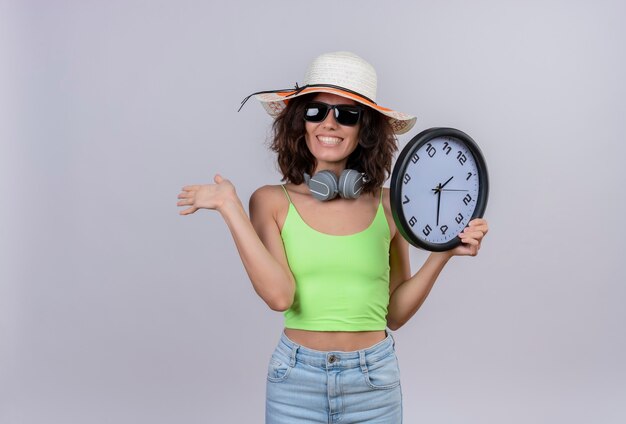 Uma jovem alegre, com cabelo curto, com top verde recortado, usando óculos escuros e chapéu de sol, segurando um relógio de parede e se despedindo em um fundo branco