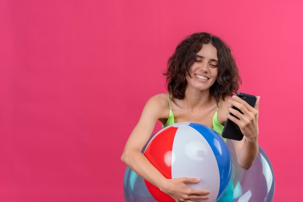 Uma jovem alegre, com cabelo curto em um top verde recortado, sorrindo e segurando uma bola inflável, olhando para o celular