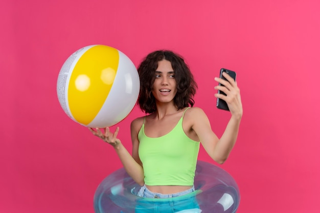 Uma jovem alegre com cabelo curto em um top verde, segurando uma bola inflável, tirando uma selfie com o celular em um fundo rosa