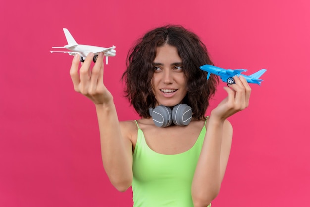 Uma jovem alegre e atraente mulher com cabelo curto e top verde recortado em fones de ouvido segurando aviões de brinquedo azuis e brancos
