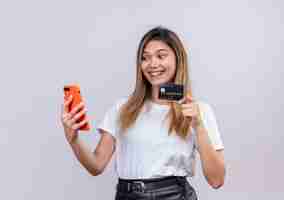 Foto grátis uma jovem alegre em uma camiseta branca mostrando o cartão de crédito enquanto olha para o celular em uma parede branca