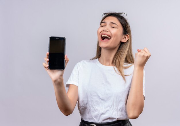 Uma jovem alegre em uma camiseta branca usando óculos escuros sorrindo enquanto mostra o espaço em branco do celular com o punho cerrado em uma parede branca