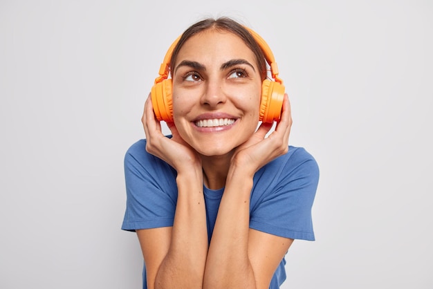 Foto grátis uma jovem alegre sonhadora mantém as mãos em sorrisos de fones de ouvido sem fio gosta agradavelmente de sua música favorita vestida com camiseta azul casual, isolada sobre fundo branco. batida de música para clima energético