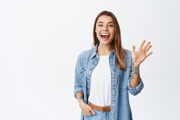 Uma jovem amigável diz oi e acena com a mão alegre sorrindo para a câmera para cumprimentá-lo em pé contra o fundo branco