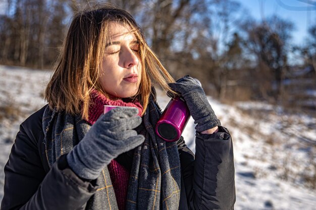 Uma jovem aprecia uma bebida quente em uma garrafa térmica em uma caminhada no inverno