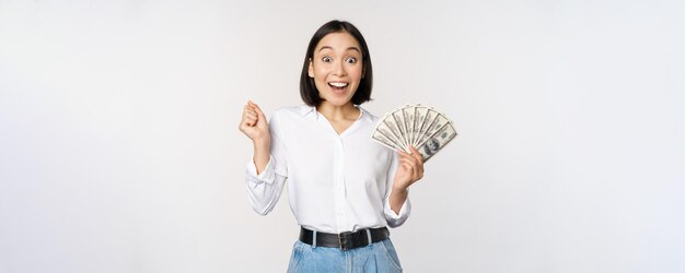 Uma jovem asiática entusiasmada olhando animada para a câmera segurando dólares de dinheiro na mão sobre fundo branco