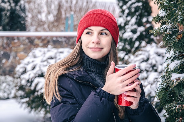 Uma jovem bebe uma bebida quente em um copo térmico vermelho no inverno