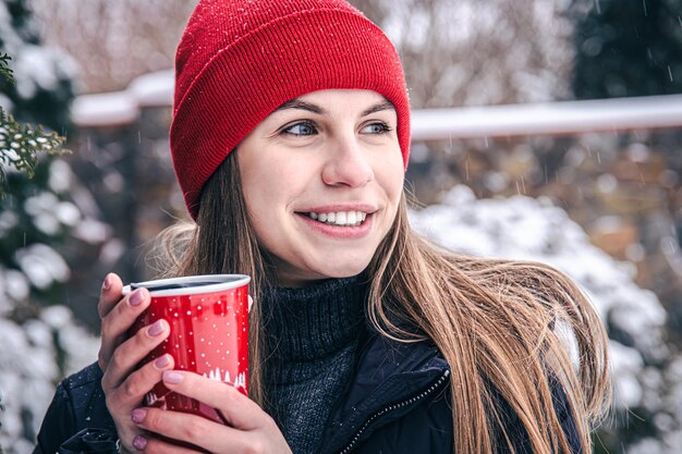 Uma jovem bebe uma bebida quente em um copo térmico vermelho no inverno