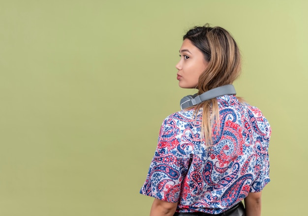 Uma jovem bonita com cabelo comprido em uma camisa estampada de paisley usando fones de ouvido, olhando para longe em uma parede verde