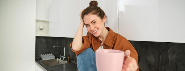 Foto grátis uma jovem bonita e sorridente senta-se na cozinha, oferece-lhe uma chávena de café, estende a mão com uma caneca rosa.