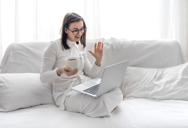 Uma jovem bonita está sentada em casa em um sofá branco com um vestido branco na frente de um laptop. Trabalho remoto e freelancer.