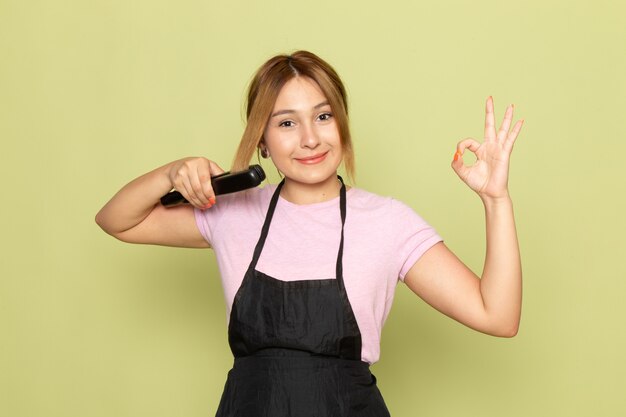 Uma jovem cabeleireira feminina de camiseta rosa e capa preta ajeitando o cabelo com uma escova de cabelo, sorrindo no verde