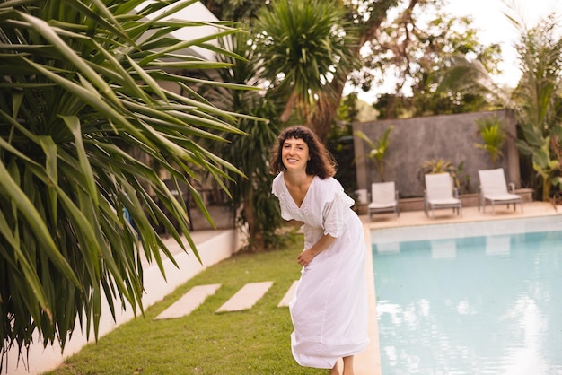 Uma jovem caucasiana engraçada com cabelo escuro em vestido branco passa tempo de férias na piscina ao ar livre