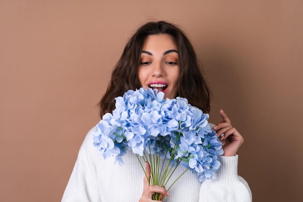 Uma jovem com cabelo volumoso ondulado em um fundo bege com brilho labial de batom rosa brilhante em um suéter branco segura um buquê de flores azuis