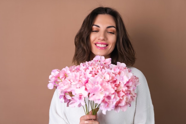 Uma jovem com cabelo volumoso ondulado em um fundo bege com brilho labial de batom rosa brilhante em um suéter branco segura um buquê de flores cor de rosa