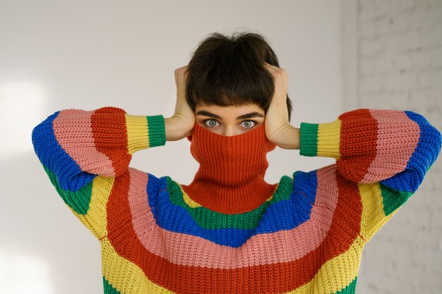 Uma jovem com um suéter de arco-íris multicolorido brilhante esconde o rosto e cobre as orelhas com as mãos.