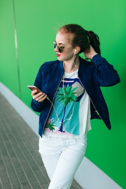 Foto grátis uma jovem com uma jaqueta azul e calça branca está em pé ao ar livre perto de uma parede verde com linha branca para baixo. a garota usa óculos escuros com corações. ela está diminuindo a música com a ajuda de fones de ouvido.