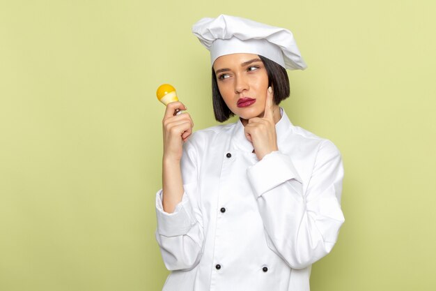 Uma jovem cozinheira de frente para o cozinheiro com um terno branco e boné segurando uma lâmpada amarela com expressão de pensamento na parede verde. senhora trabalhar comida culinária cor