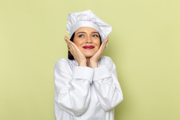 Uma jovem cozinheira de frente para um cozinheiro com um terno branco e um boné posando com uma expressão encantada na parede verde.