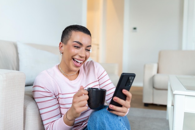 Uma jovem de suéter com uma xícara nas mãos olha para o telefone enquanto está sentada no sofá