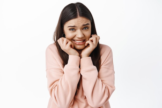 Uma jovem desajeitada se sente desconfortável apertando os dentes e parecendo culpada, preocupada em ficar ansiosa ou nervosa contra um fundo branco usando capuz