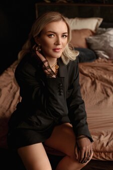 Uma jovem deslumbrante com cabelos loiros e um vestido preto posa na cama no interior luxuoso e escuro