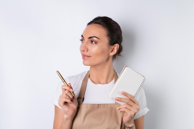 Uma jovem dona de casa de avental em um fundo branco com um caderno nas mãos escolhe receitas para o jantar pensa em como agradar o marido tem ideias para pratos