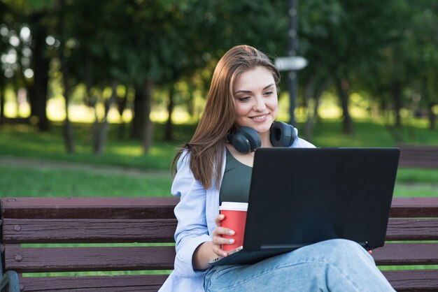 Uma jovem e atraente garota caucasiana em fones de ouvido, sentado em um banco do parque. usando um laptop. freelance, conceito de rede