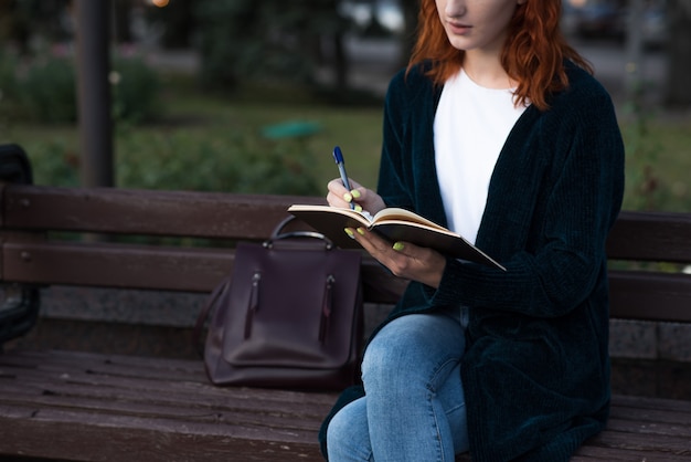 Uma jovem e atraente ruiva caucasiana, escrevendo em um caderno enquanto está sentado em um banco. conceito de educação.