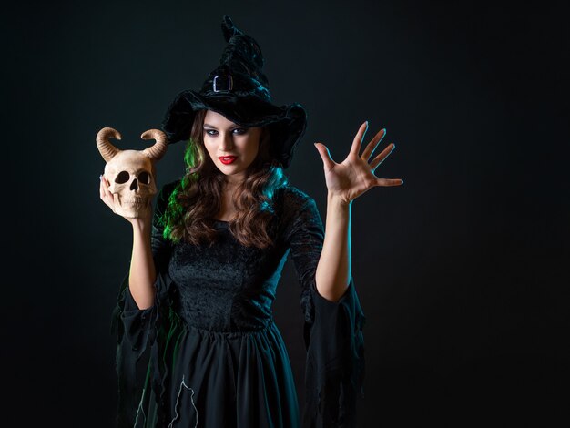 Uma jovem e bela bruxa com um chapéu cônico segura uma caveira do diabo nas mãos