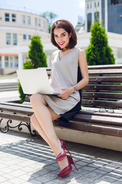 Uma jovem empresária linda morena está sentada no banco na cidade. Ela usa vestido cinza e preto e salto alto. Ela está digitando no laptop e sorrindo para a câmera.