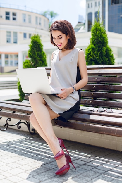 Uma jovem empresária linda morena está sentada no banco na cidade. Ela usa vestido cinza e preto e salto alto. Ela está digitando no laptop.