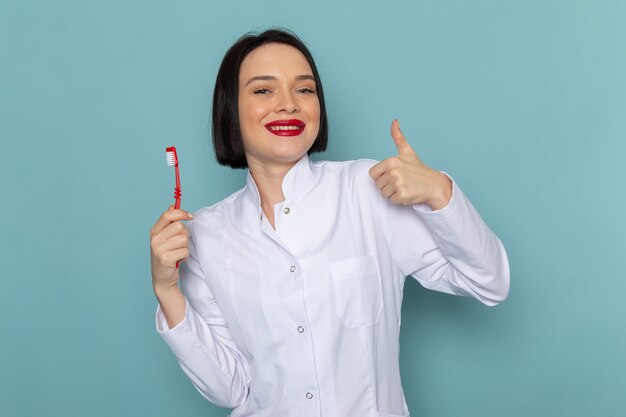 Uma jovem enfermeira com terno branco e sorrindo, segurando a escova de dentes na mesa azul.